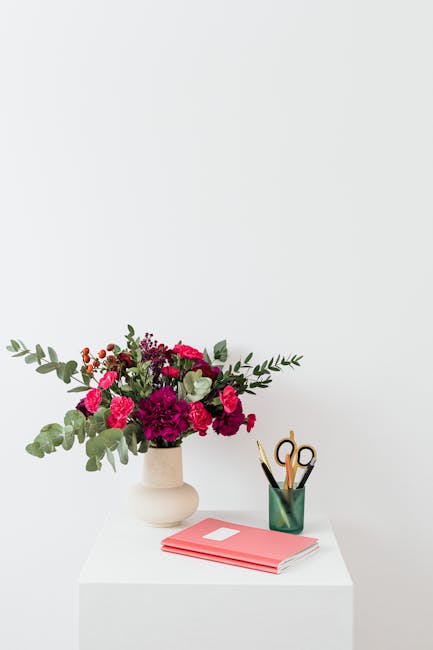 A fresh floral arrangement in a white ceramic vase featuring deep magenta and bright pink carnations, accented with green eucalyptus and other foliage, is placed on a white table against a plain light