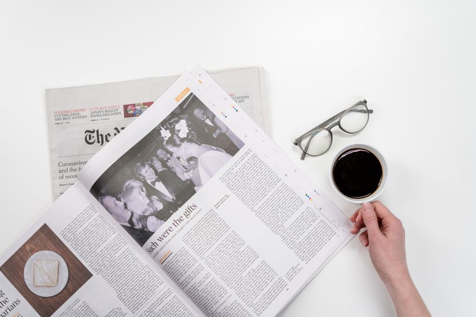 A person holding a white ceramic mug filled with black coffee, placed on a white surface alongside an open newspaper with a black-and-white photo and text, a pair of reading glasses, and a section of 