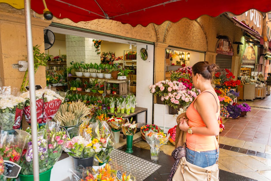 A woman in a peach sleeveless top and blue jeans stands outdoors in front of a flower shop, [COMPANY_NAME], on Kensington High Street, browsing a colorful floral display. The arrangement includes pink