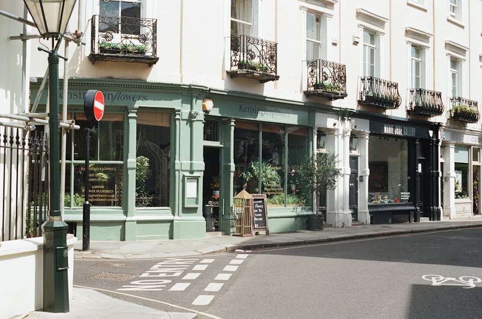 A view of a flower shop on Kensington High Street, with a mint green storefront showcasing large glass windows filled with floral displays. The shop, associated with floristwestkensington.co.uk, featu