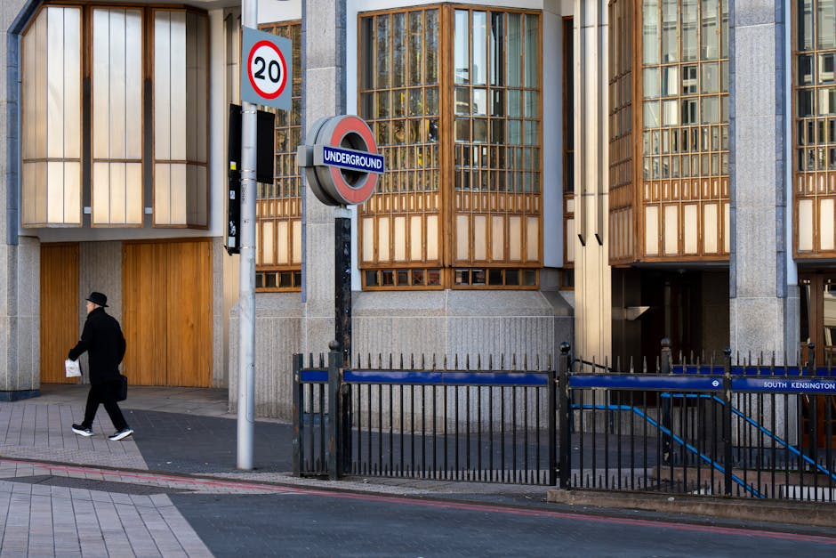 The image shows a scene outside a modern building with large wooden-framed windows, located near Olympia Station in Kensington. A man dressed in black, wearing a hat, is walking along the sidewalk car