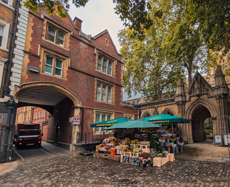 A street scene in Kensington featuring a florist's stall under a green canopy, displaying a vibrant array of fresh flowers including pink roses, white lilies, and yellow tulips arranged in wooden crat