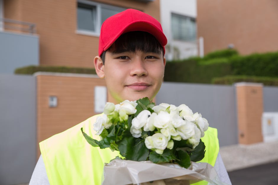 A young male flower delivery courier wearing a red cap and high-visibility yellow vest is holding a bouquet of white roses wrapped in clear and white paper, smiling softly. The arrangement features fr