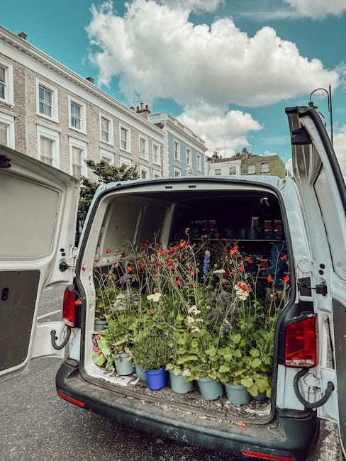 The open rear of a white delivery van filled with potted flowers, including tall red poppies, white daisies, and other green foliage, arranged in blue and gray pots. The flowers appear fresh with vibr