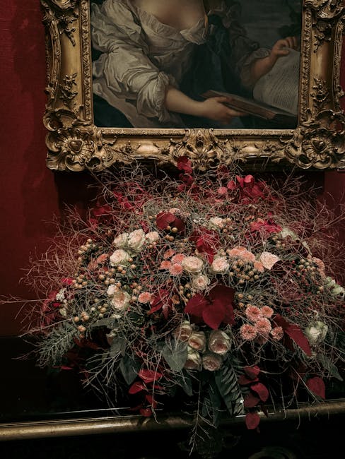 A large, lush floral arrangement featuring a variety of flowers in shades of pink, white, and deep red, including roses and smaller filler blooms, accented with dark green foliage and wispy, dried bra