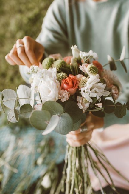 A close-up of a person holding a floral bouquet featuring a mix of white, peach, and soft pink roses, eucalyptus leaves, and delicate white flowers, arranged in a natural, loosely bound style with vis