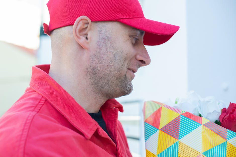 A delivery person wearing a red cap and matching red jacket is holding a bouquet of fresh flowers wrapped in white tissue paper and colorful geometric-patterned paper. The bouquet features a variety o