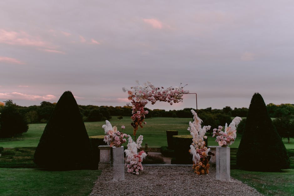 A floral arrangement consisting of pink and white flowers, including roses, peonies, and delphiniums, artfully displayed on white marble pedestals, with tall, cone-shaped evergreen shrubs on either si