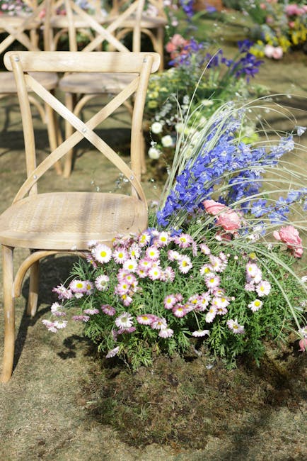 A floral arrangement featuring a mix of pink roses, purple delphiniums, and white chrysanthemums, planted in a rustic bed of earth. The arrangement is complemented by tall, wispy grasses and green fol