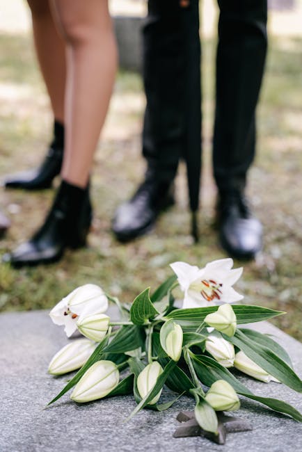 A bouquet of fresh white lilies with elegant, large blossoms and elongated green leaves rests on a stone surface. Surrounding the lilies are several unopened buds, showcasing a smooth, creamy petal te