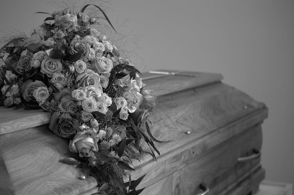 A bouquet of fresh flowers rests on a wooden casket, featuring predominantly roses in various stages of bloom, with some fully open and others still budding. The flowers are in shades of light and dar